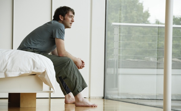 Young man sitting on edge of bed, looking out patio doors, side view